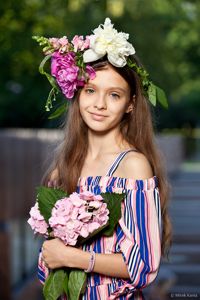 Zdjęcie przedstawia młodą dziewczynę w wianku, trzymającą w rękach kwiaty. W tle widzimy amfiteatr SDK.