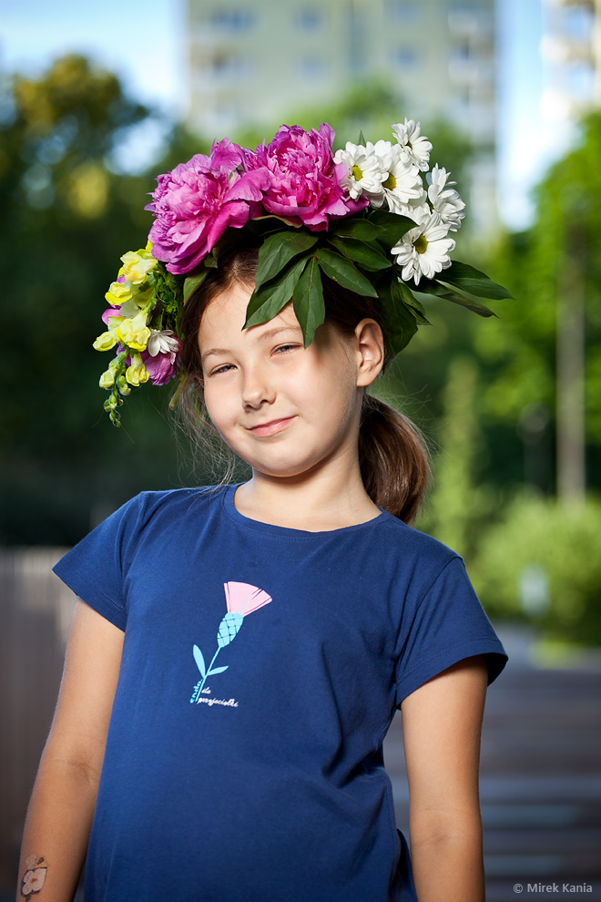 Na zdjęciu pokazana jest młoda dziewczynka w wianku. W tle widzimy drzewa oraz fragment bloku mieszkalnego.