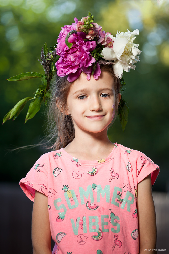 Na zdjęciu widoczna jest młoda dziewczynka w wianku. W tle widoczne są drzewa.