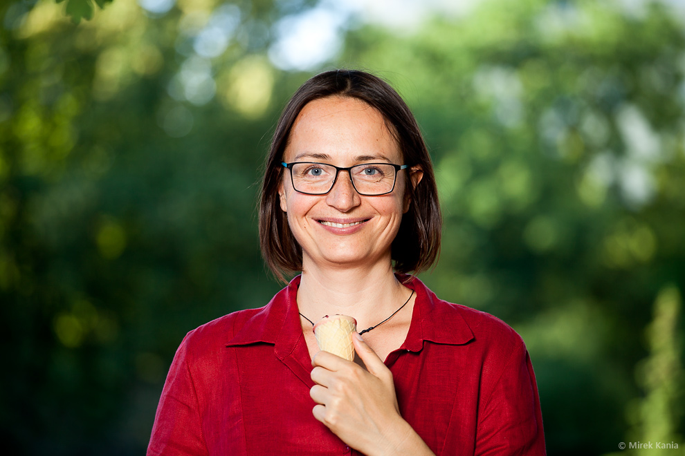 Na zdjęciu widoczna jest kobieta z lodem w ręku. W tle widać zieleń drzew. 