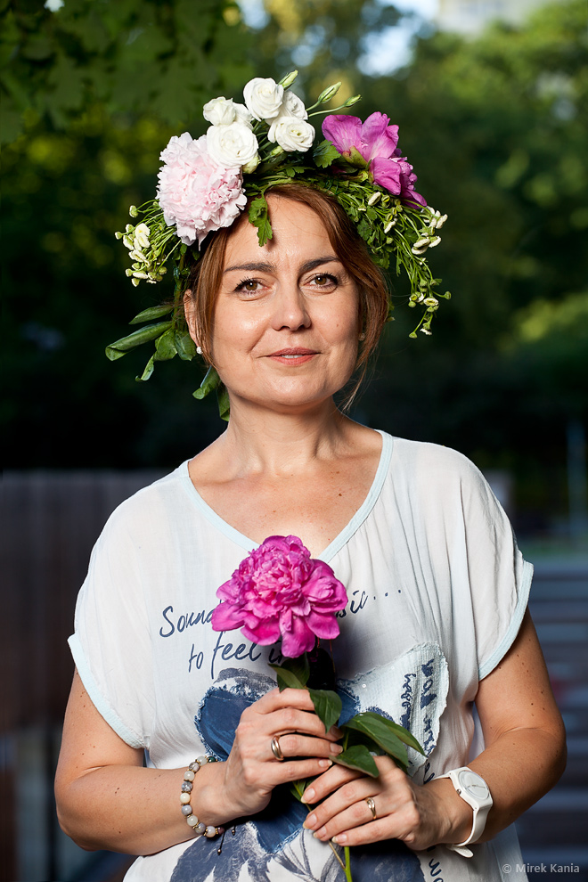 Na zdjęciu widoczna jest kobieta w wianku z kwiatem w rękach. W tle widoczny jest amfiteatr SDK oraz zieleń parku.