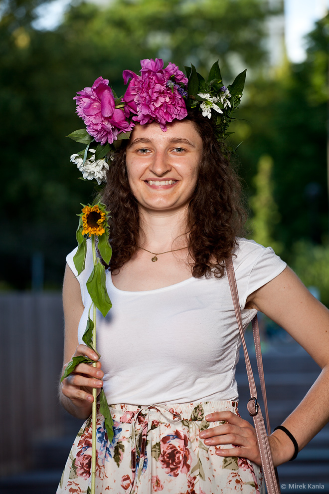 Na zdjęciu widoczna jest młoda kobieta w wianku trzymająca kwiat w dłoni. W tle widać amfiteatr SDK.