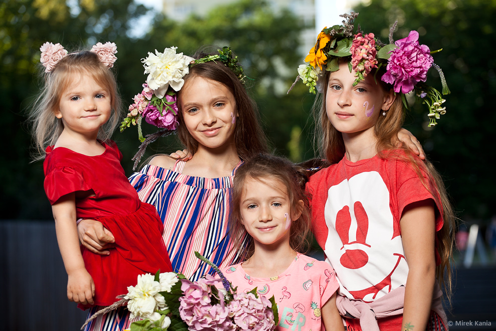 NA zdjęciu widoczne są cztery młode dziewczynki, najmłodsza trzymana jest na rękach. Trzy dziewczynki mają na głowie wianki, czwarta trzyma w ręku kwiat. W tle zieleń parku.