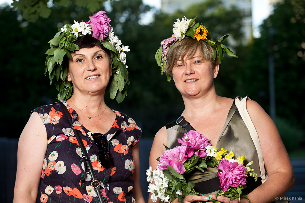 Zdjęcie przedstawia dwie kobiety we wiankach. Jedna z nich trzyma bukiet kwiatów w rękach. W tle widać park SDK.