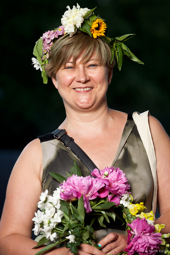 Zdjęcie przedstawia kobietę w wianku z bukietem kwiatów w rękach. Tło ciemne, przedstawia park. 