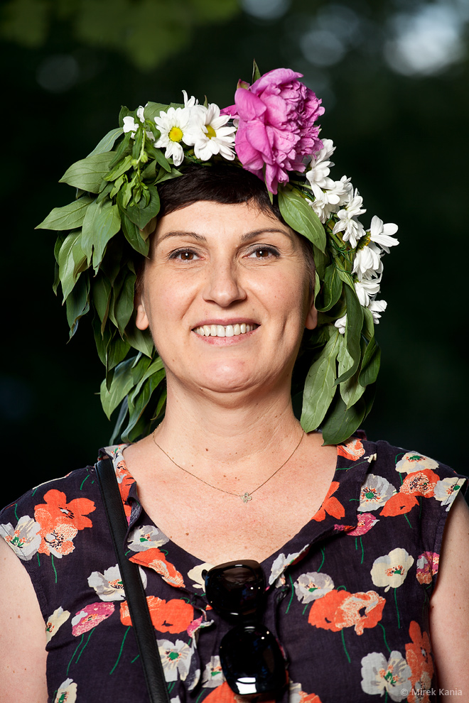 Zdjęcie portretowe prezentuje uśmiechniętą kobietę w wianku. W tle widać zieleń drzew. 