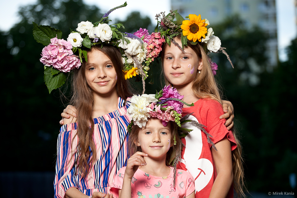 Zdjęcie przedstawia trzy młode dziewczynki w wiankach. W tle widać blok mieszkalny oraz zieleń drzew. 