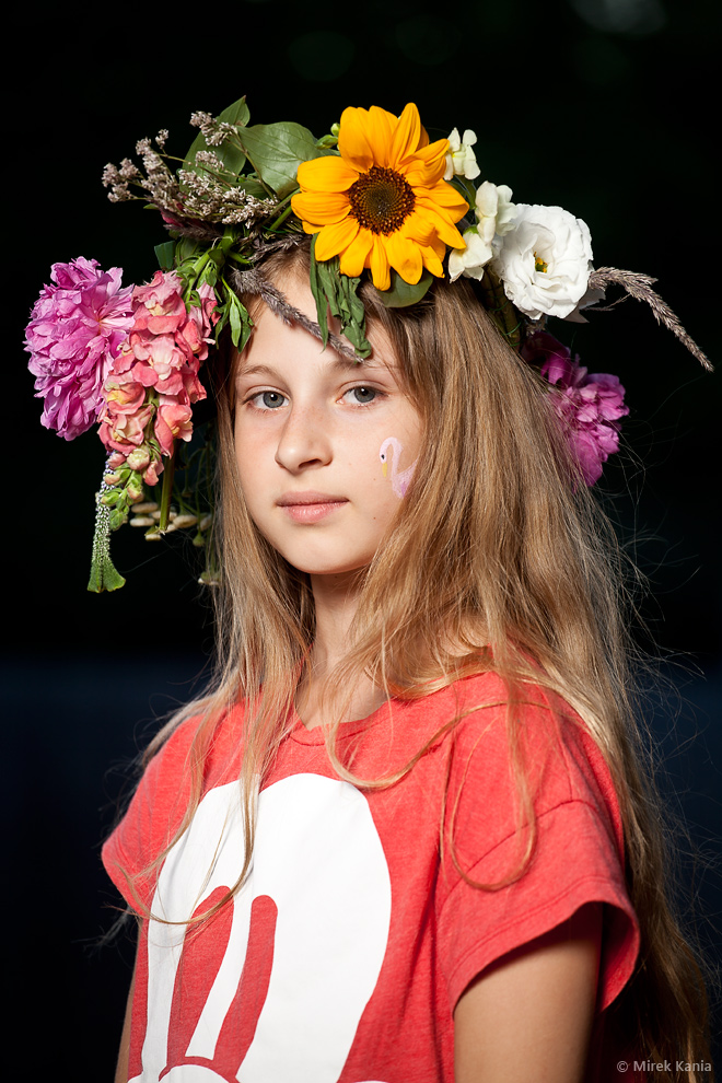 Zdjęcie portretowe prezentuje dziewczynkę w wianku na ciemnym tle.  