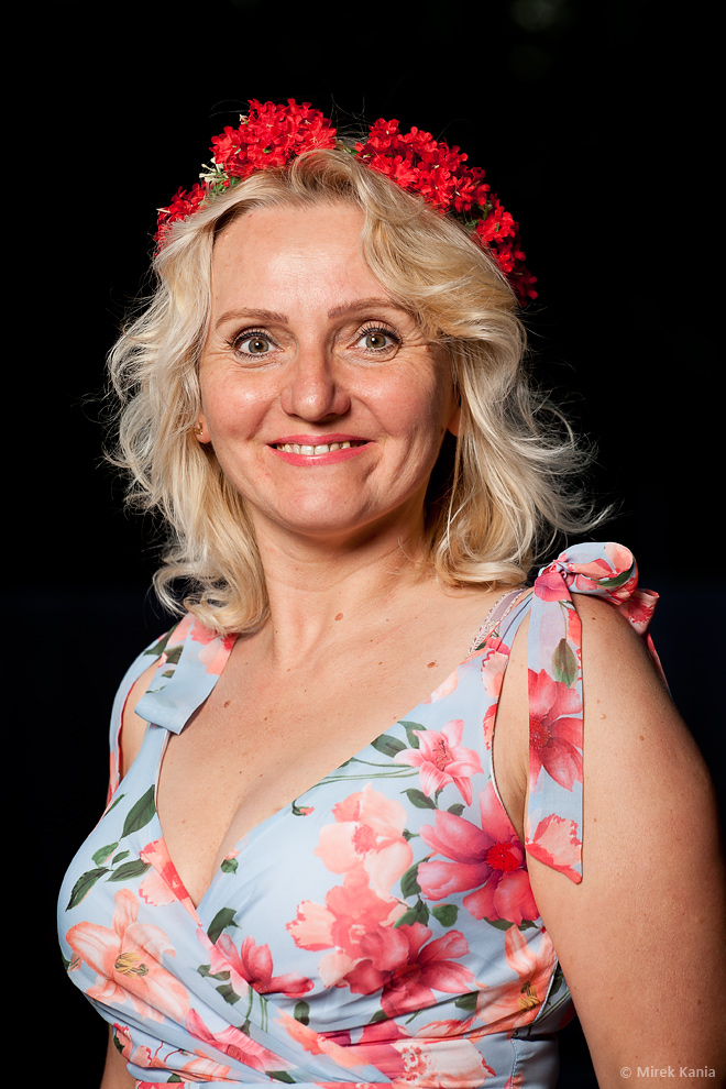 Zdjęcie przedstawia kobietę w wianku na ciemnym tle. Kobieta ubrana jest w kwiecistą sukienkę. 