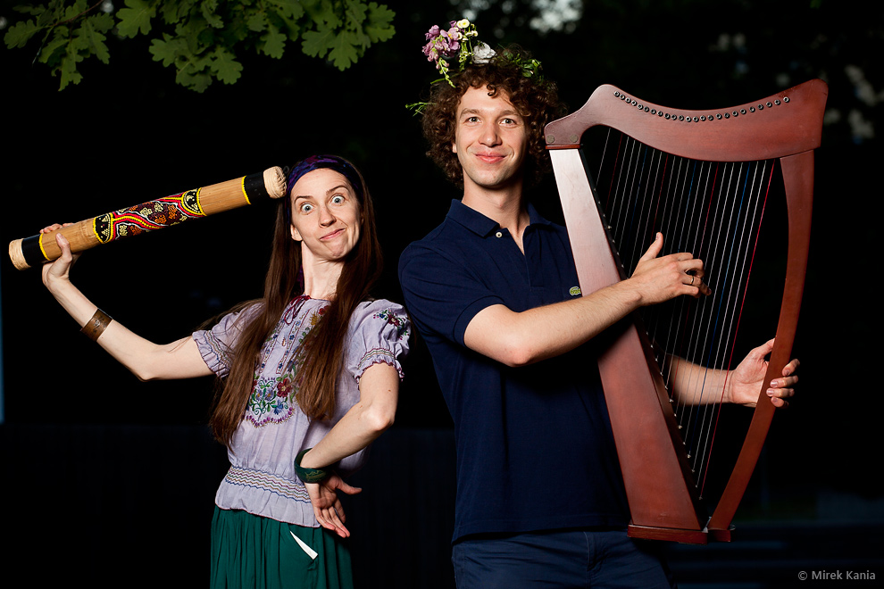 Zdjęcie przedstawia dwie młode osoby. Mężczyzna trzyma instrument przypominający harfę, kobieta drewnianą tubę. Oboje przyjęli zabawne pozy i miny. 