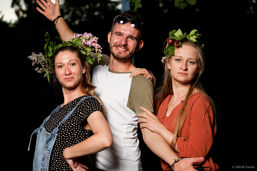 Zdjęcie przedstawia trzy osoby. Dwie młode kobiety oraz młodego mężczyznę. Kobiety we wiankach, mężczyzna ma rozłożone ręce w geście powitania. Ciemne tło. 