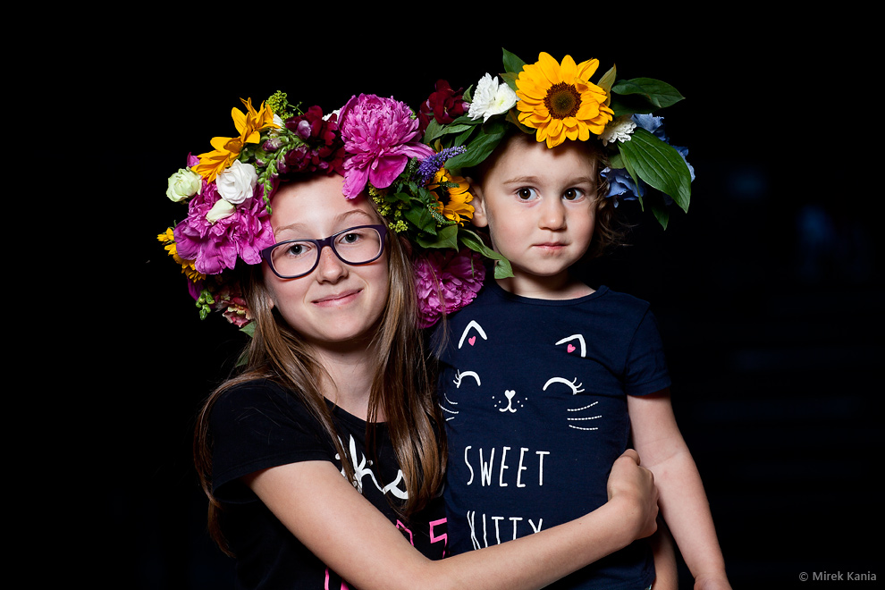 Zdjęcie przedstawia młodą dziewczynę w wianku trzymającą na rękach młodszą dziewczynkę. Ciemne tło. 