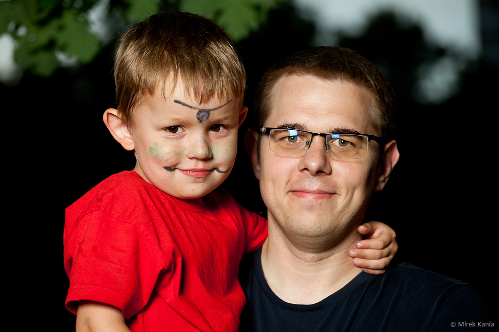 Zdjęcie przedstawia mężczyznę z małym chłopcem w czerwonej koszulce na rękach. Za nimi ciemne tło. 