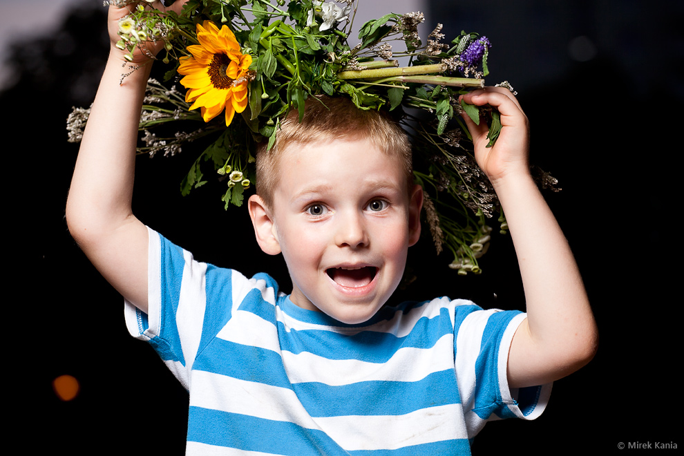 Na zdjęciu widać małego chłopca w wianku na głowie w dynamicznej pozie na ciemnym tle. 