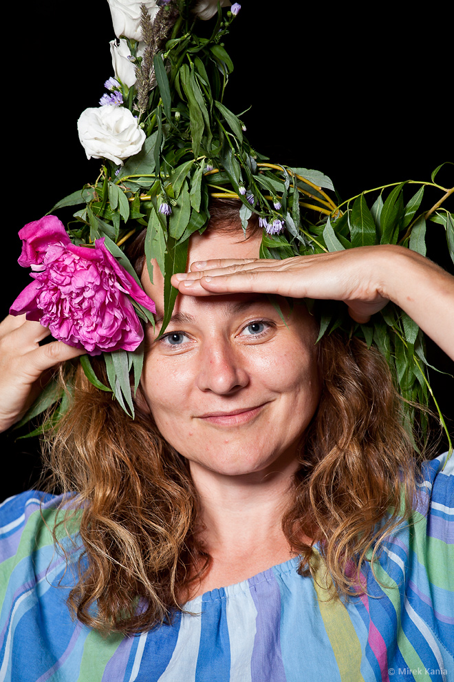 Na zdjęciu widoczna jest kobieta w wianku trzymająca rękę jakby patrzyła w dal. Ciemne tło.