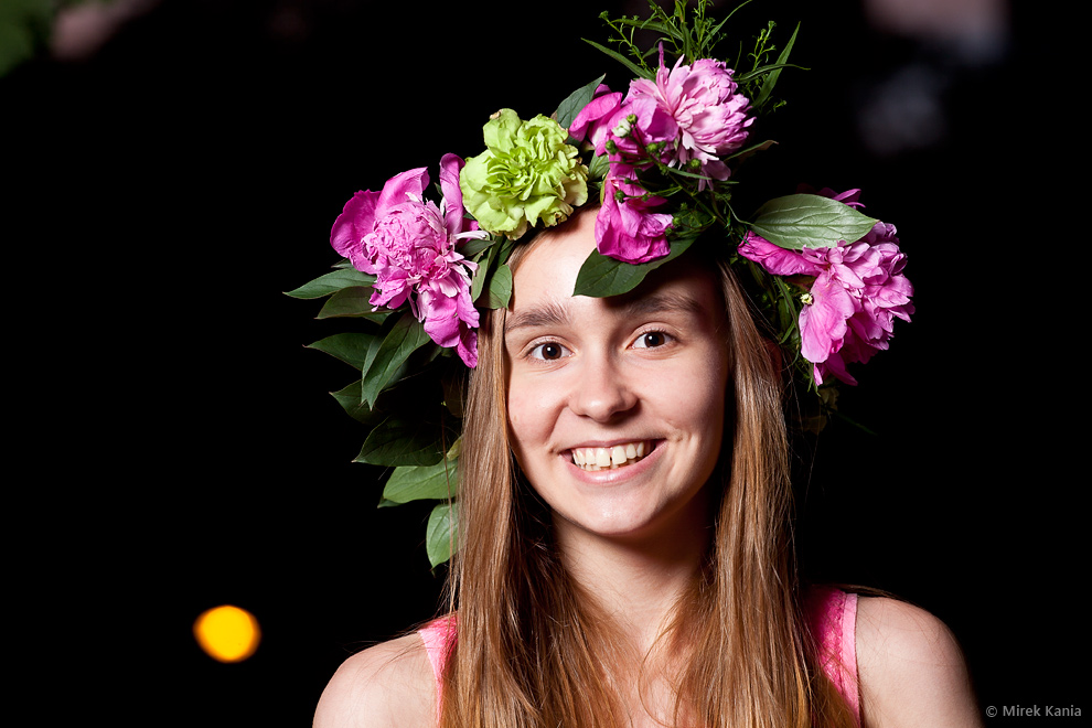 Zdjęcie pokazuje młodą uśmiechniętą dziewczynę w wianku. Ciemne tło. 