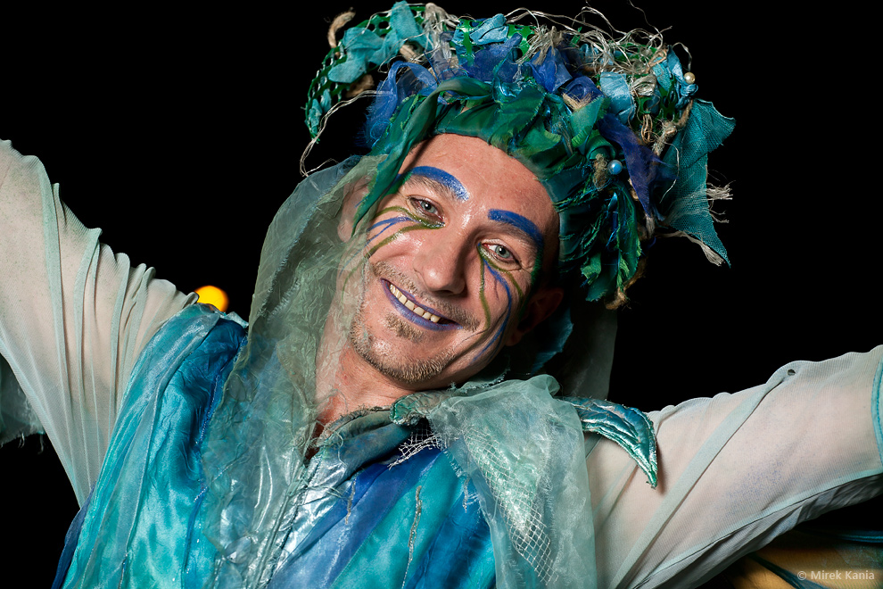 Zdjęcie przedstawia aktora spektaklu ubranego w niebiesko - białe ubranie, w makijażu oraz czapce ze strzępków materiału. Mężczyzna ma rozłożone szeroko ręce. Ciemne tło.