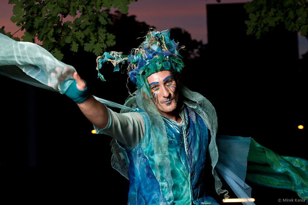 Zdjęcie prezentuje aktora w niebieskim ubraniu z makijażem w czapce ze strzępków materiału. Mężczyzna wyciąga rękę w stronę aparatu. Ciemne, nocne tło. 