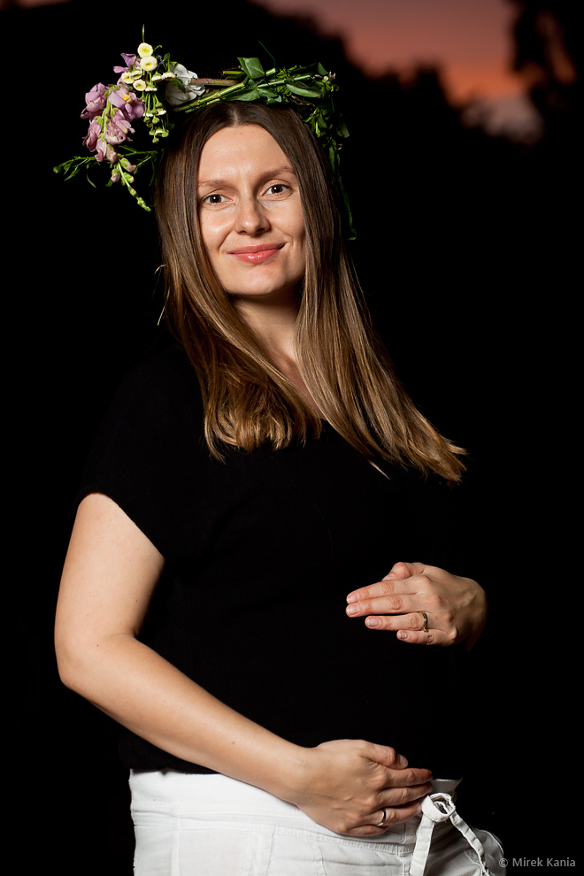 Zdjęcie przedstawia kobietę w ciąży w wianku na głowie. Kobieta trzyma się za brzuch. Ciemne, nocne tło.