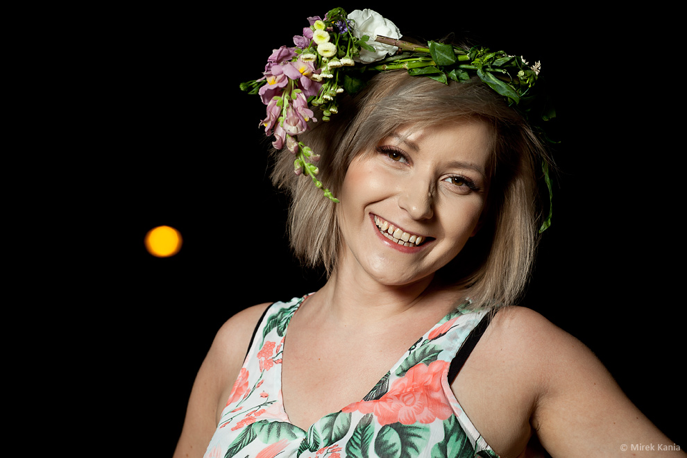 Zdjęcie przedstawia młodą kobietę w wianku. Ciemne tło. 