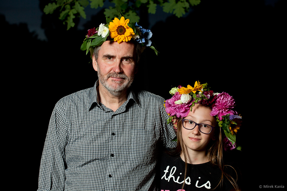 Na zdjęciu widoczny jest mężczyzna w średnim wieku obejmujący młodą dziewczynkę. Obydwoje w wiankach na głowie. Ciemne tło.