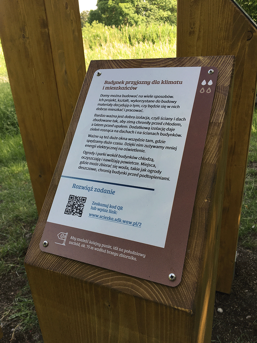 Tablica projektu Klimatycznej ścieżki edukacyjnej Zdjęcie prezentuje tabliczkę informacyjną ścieżki. W tle widoczny Park Służewskiego Domu Kultury.