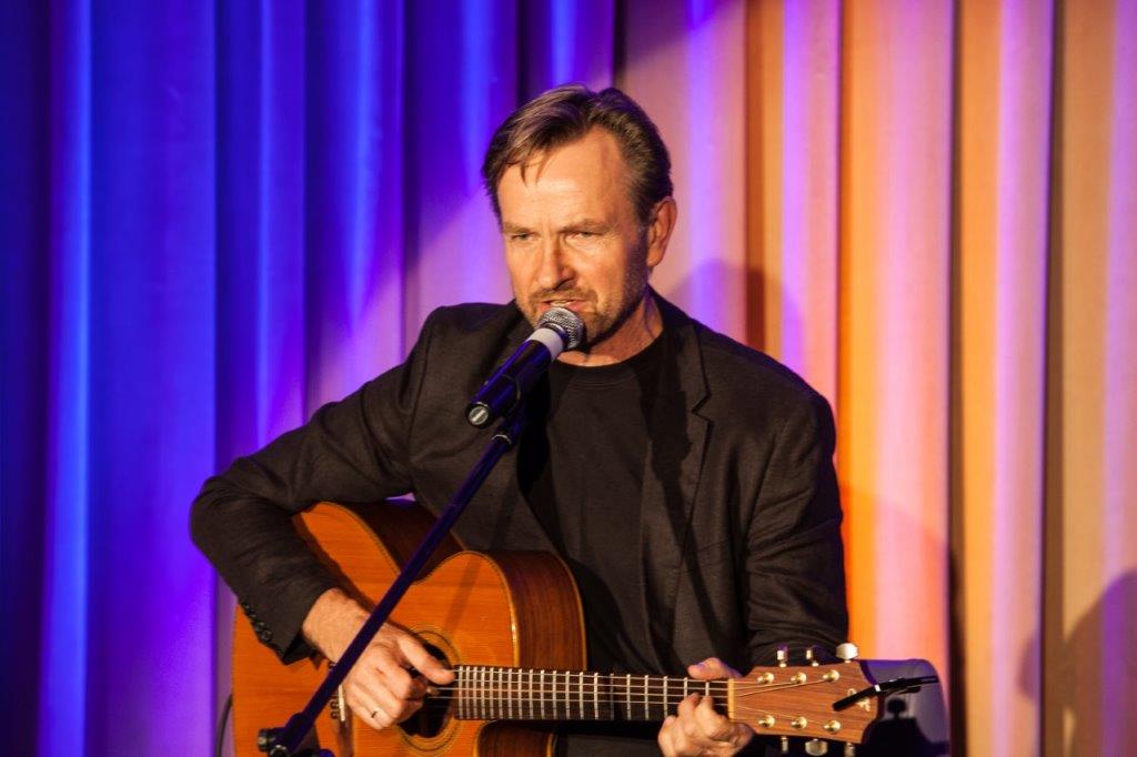 Grafika przedstawia Artystę podczas koncertu. Trzyma w rękach gitarę, w tle kurtyna oświetlona światłem scenicznym