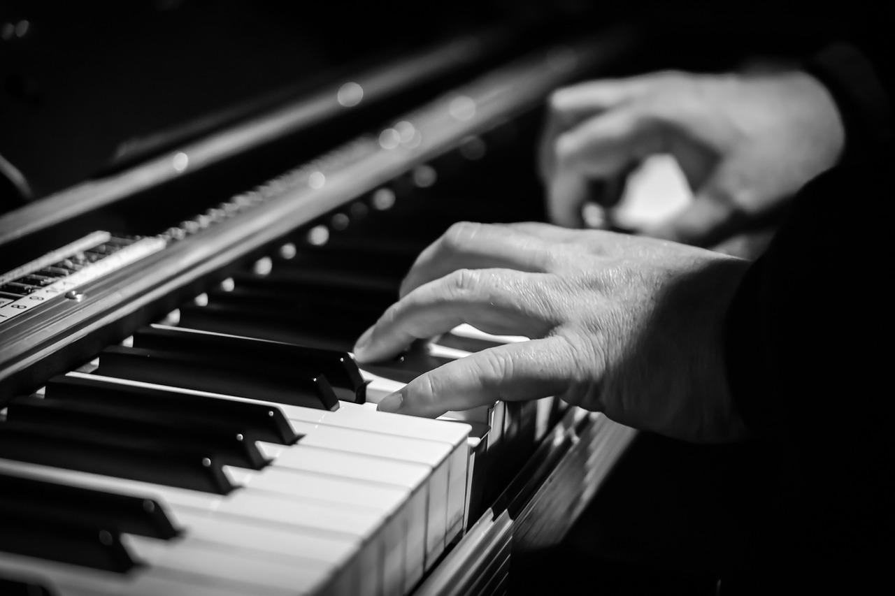 Monochromatyczna fotografia prezentująca zbliżenie na dłonie pianisty grające na klawiaturze fortepianowej.