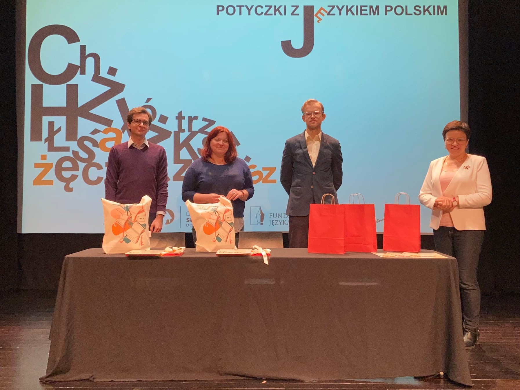 Na zdjęciu: wszyscy jurorzy dyktanda stojący na scenie przy stole z nagrodami. Za nimi ekran z grafiką identyfikującą wydarzenie.