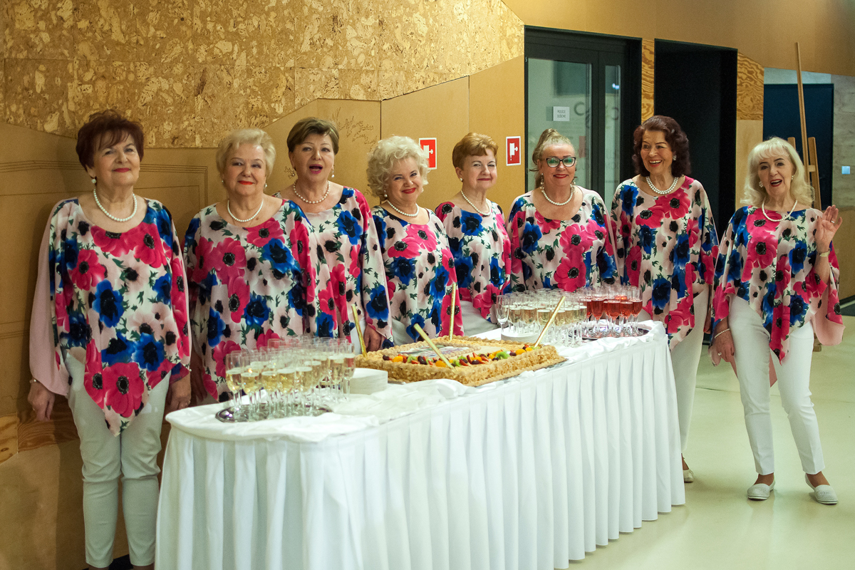 Na zdjęciu osiem pań - wokalistek przed urodzinowym tortem ustawionym na białym podłużnym stole. Paniew ubrane są w białe spodnie i kwieciste bluzki. W tle korkowa ściana.