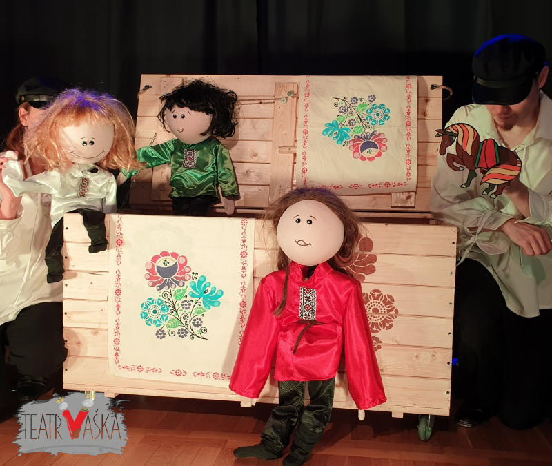 Na zdjęciu dwójka aktorów trzyma w rękach lalki. Obok nich dwie kolejne lalki - jedna siedzi na drewnianej skrzyni, a druga stoi na ziemii. Wszystkie postaci znajdują się na scenie. W tle kurtyna.