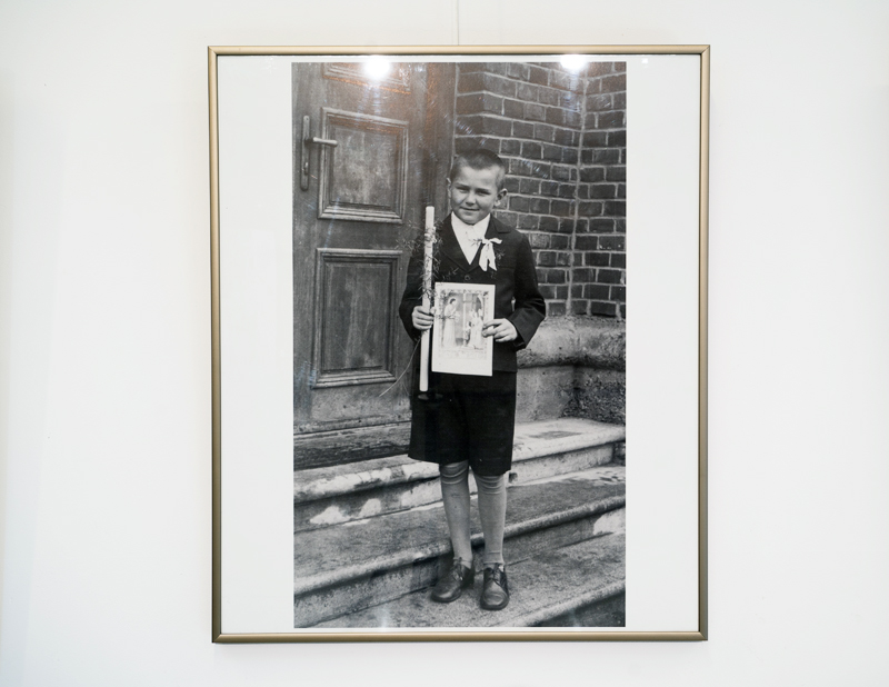 Pamiątkowe zdjęcie z Pierwszej Komunii Św. stojącego na schodzach kościoła Zdzisława Beksińskiego, trzymającego świecę oraz pamiątkowy obrazek z komunii.