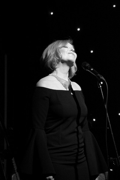 Na zdjęciu: Justyna Bacz na scenie przy mikrofonie; ciemne jednolite tło i jasna kontrastowa twarz artystki. Autor fot. . J. Szlapka. Fotografia wykonana  w Teatr Kamienica podczas "Dalida Pieśń Miłości" - 31.03 2017 r. Zdjęcie ze strony http://www.justynabacz.net/