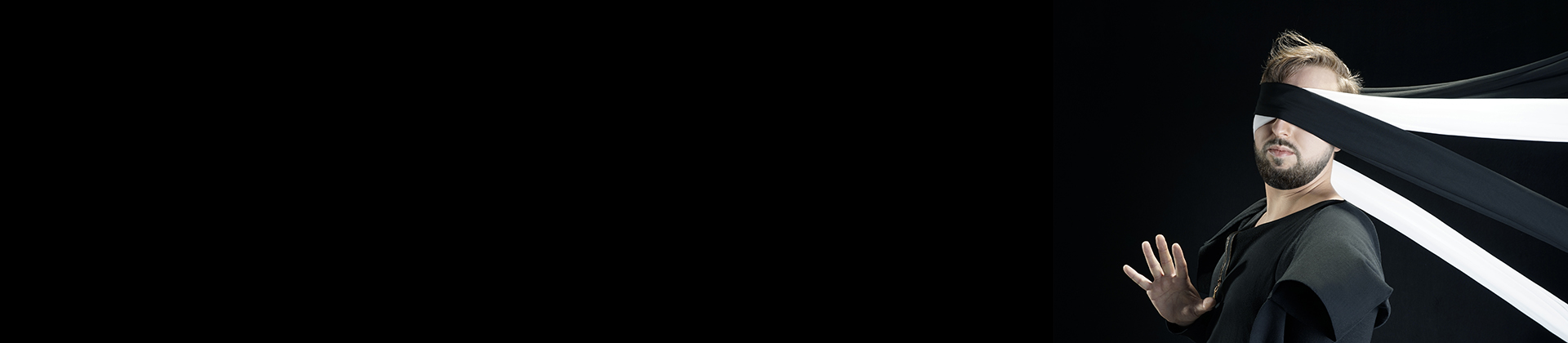 Na zdjęciu czarne tło, po prawej stronie grafiki artystya o blond włosach, w ciemnym ubraniu na czarnym tle ma zasłonięte oczy dwiema długimi tasmami w kolorze czerni i bieli.