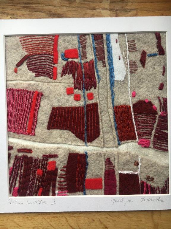 Zdjęcie pzredstawia pracę - mieszaninę technik z zastosowaniem tkaninowych aplikacji i haftu w wielu odcienach czerwieni. Obraz przedstawia plan miasta na lnianym tle. Obramowanie prac białe.