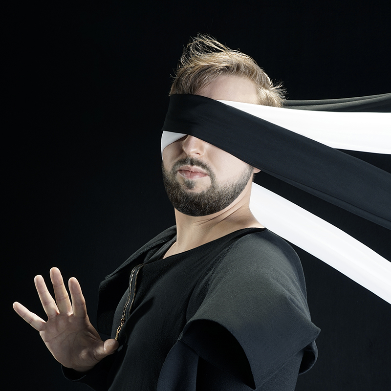 Na zdjęciu artysta o blond włosach, w ciemnym ubraniu na czarnym tle ma zasłonięte oczy dwiema długimi taśmami w kolorze czerni i bieli.