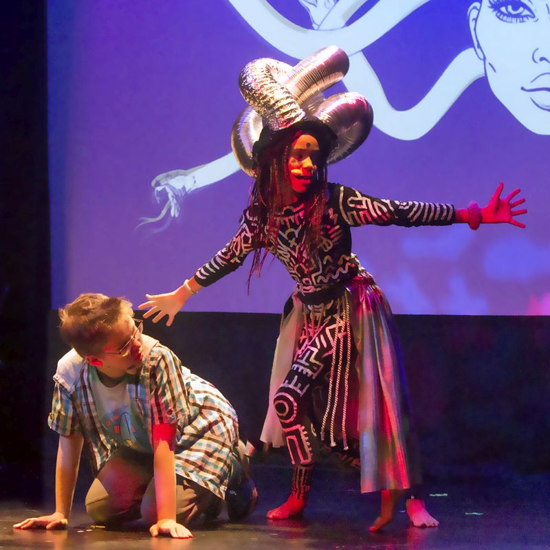 Na zdjęciu dziewczyna występująca w przedstawieniu. Stoi na froncie sceny z napiętymi dłońmi robiąc złą minę.  Przebrana za wojowniczkę stoi na scenie. W tle widoczne pozostałe osoby grające w spektaklu, osoby stoją w czerwonym świetle. 