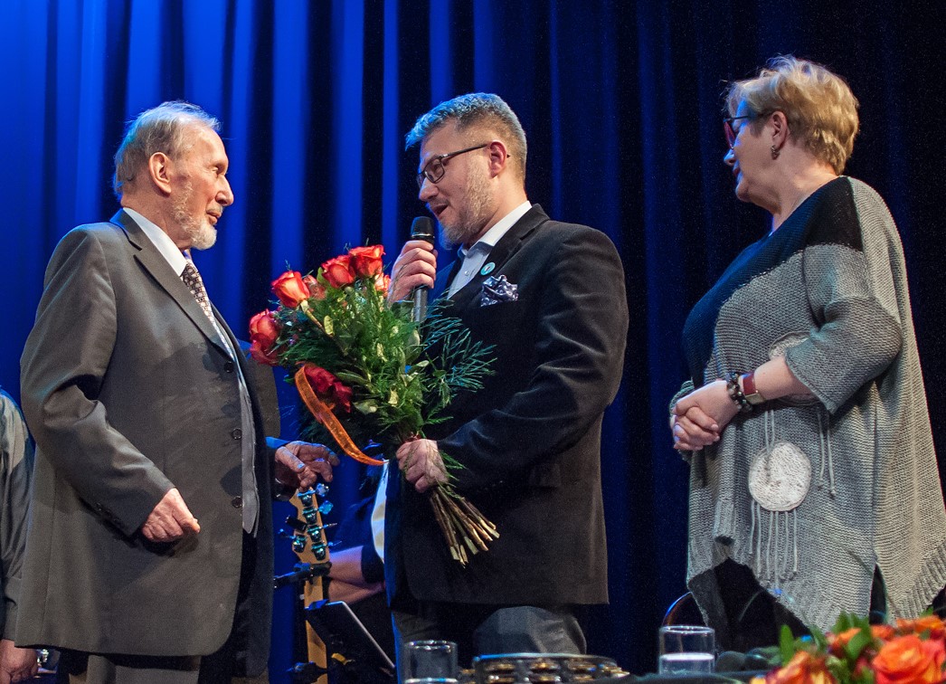 Na zdjęciu widać Jubilata i Burmistrza Mokotowa Rafała Miastowskiego. Burmistrz wręcza bukiet róż i przemawia do mikrofonu patrząc w kierunku jubilata. W tle kurtyna sceny oświetlona na niebiesko