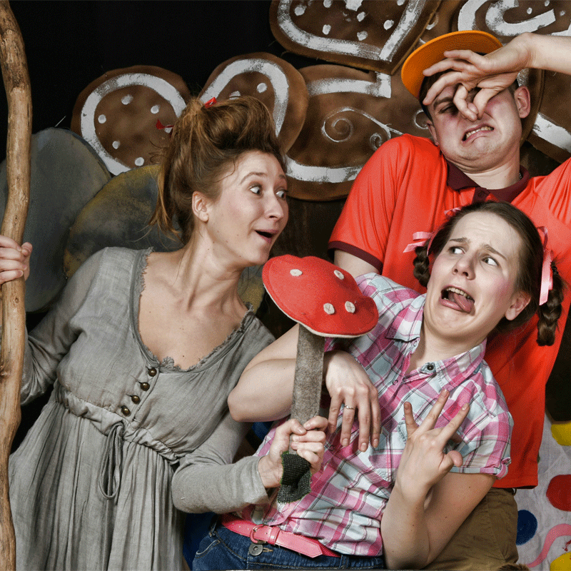 Na zdjęciu trójka aktorów, dwie kobiety i jeden mężczyzna. Aktorka grająca wiedźmę trzyma w ręku laskę oraz grzyba muchomora. Mężczyzna trzyma ręce zatykając nos. Druga kobieta odsuwa się od wiedźmy w geście obrzydzenia. 