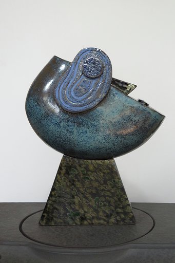 Zdjęcie przedstawia rzeźbe ceramiczną przedstawiającą artystyczną wizję głowy z okiem. Podstawa rzeźby w odcieniach szarobrązowych, część główna rzeźby w barwach niebieskich