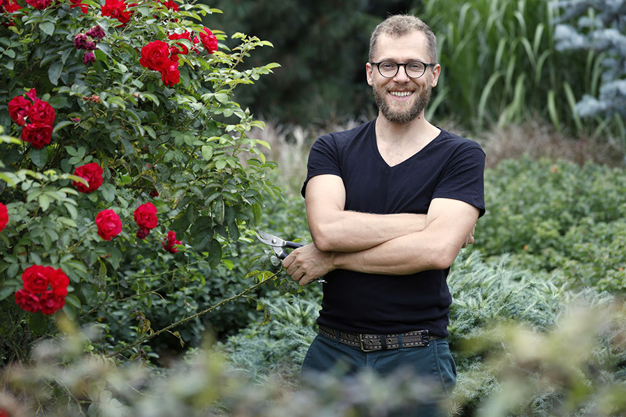 Nazdjęciu prowadzący warsztat. Stoi z sekatorem przy krzaku róż w ogrodzie. Foto: Zasoby Łukasza Szkopa