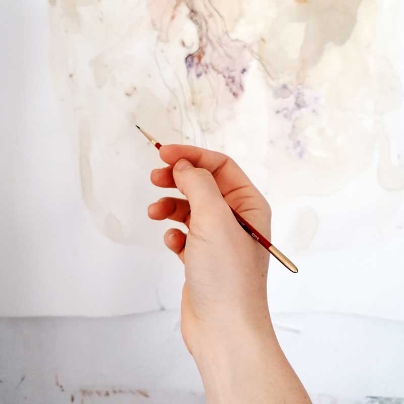 Na zdjęciu: dłoń trzymająca pędzelek na tle abstakcyjnych wzorów wykonanych barwnikiem roślinnym