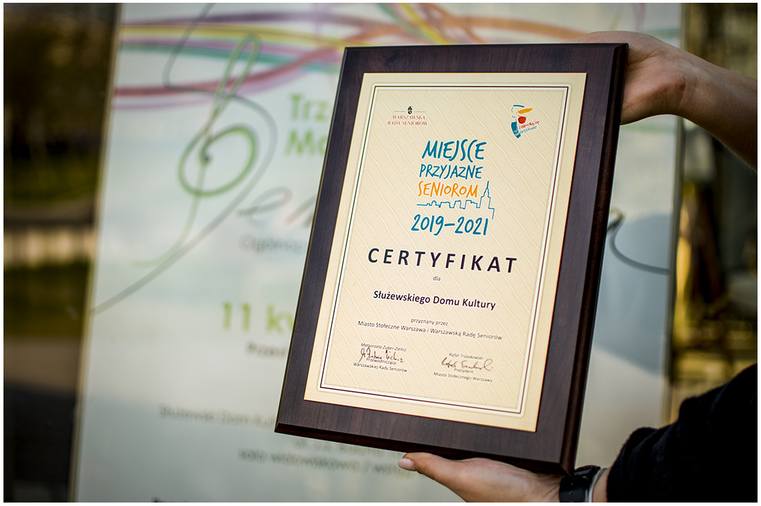 Zdjęcie promocyjne pracowni. Na zdjęciu tabliczka certyfikat dla SDK jako miejsca przyjaznego seniorom. W tle widzimy plakat reklamowy Trzecich Mokotowskich Senioraliów