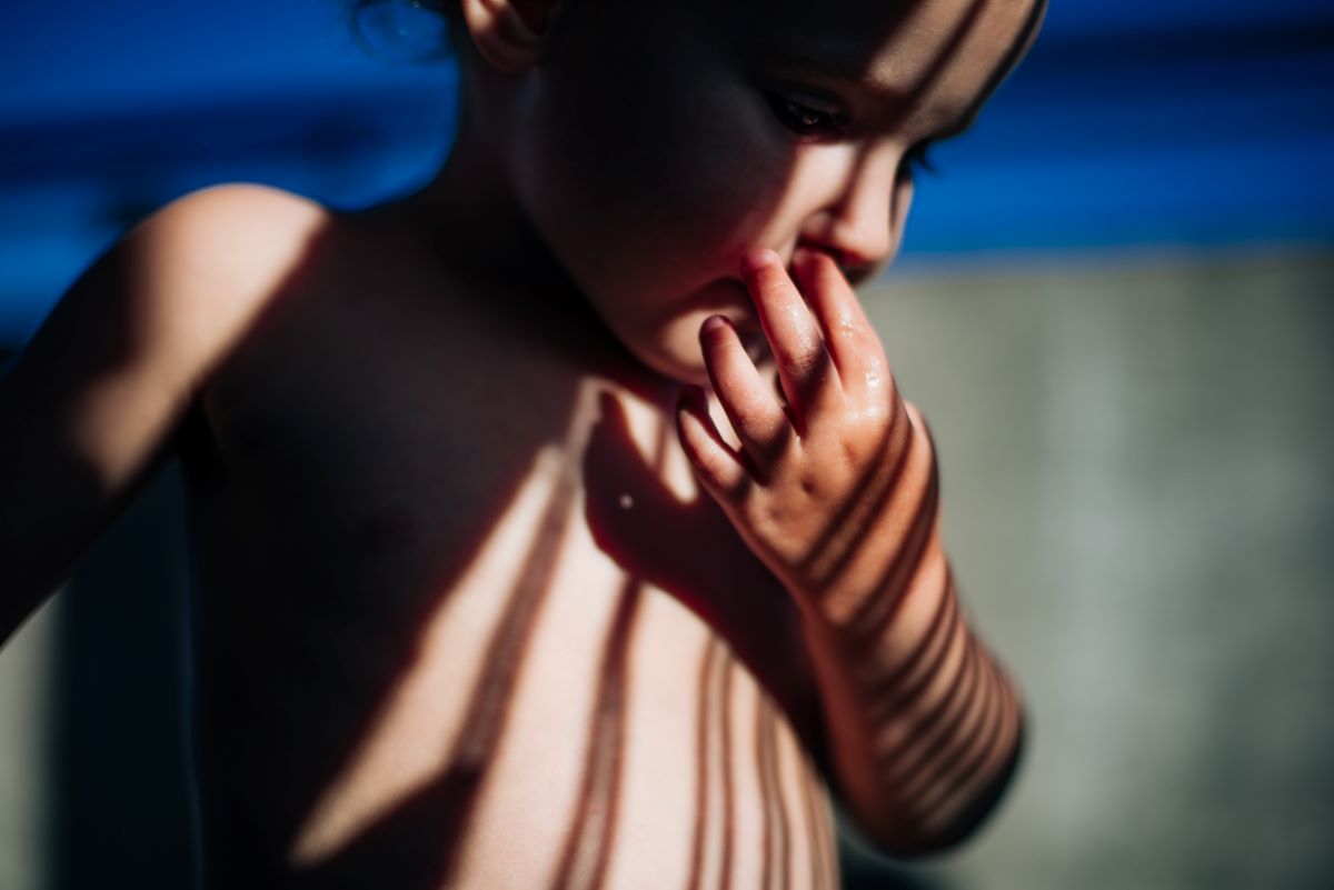 Prawie cały kadr wypełnia portret nagiego dziecka dotykającego paluszkami warg. Wzrok dziecka jest skupiony na ziemię. Na ciele dziecka widać cienie. Fot. Agnieszka Mocarska