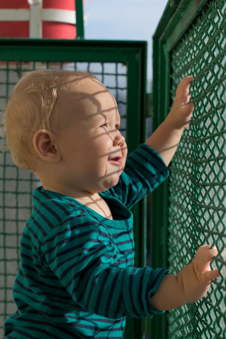 Na zdjęciu widać dziecko w zielonym body w paski na zielonym balkonie ze skrzywioną miną od promieni słońca. Fot. Zuzanna Jach