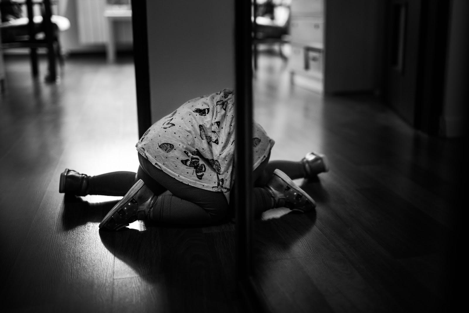 Czarno-biała fotografia. Na niej widoczny fragment dziecka wchodzącego do szafy za przesuwnymi, szklanymi drzwiami. Nie widoczna jest główka dziecka. W odbiciu lustra widoczne są nóżki dziecka. W tle widoczny fragment mieszkania.