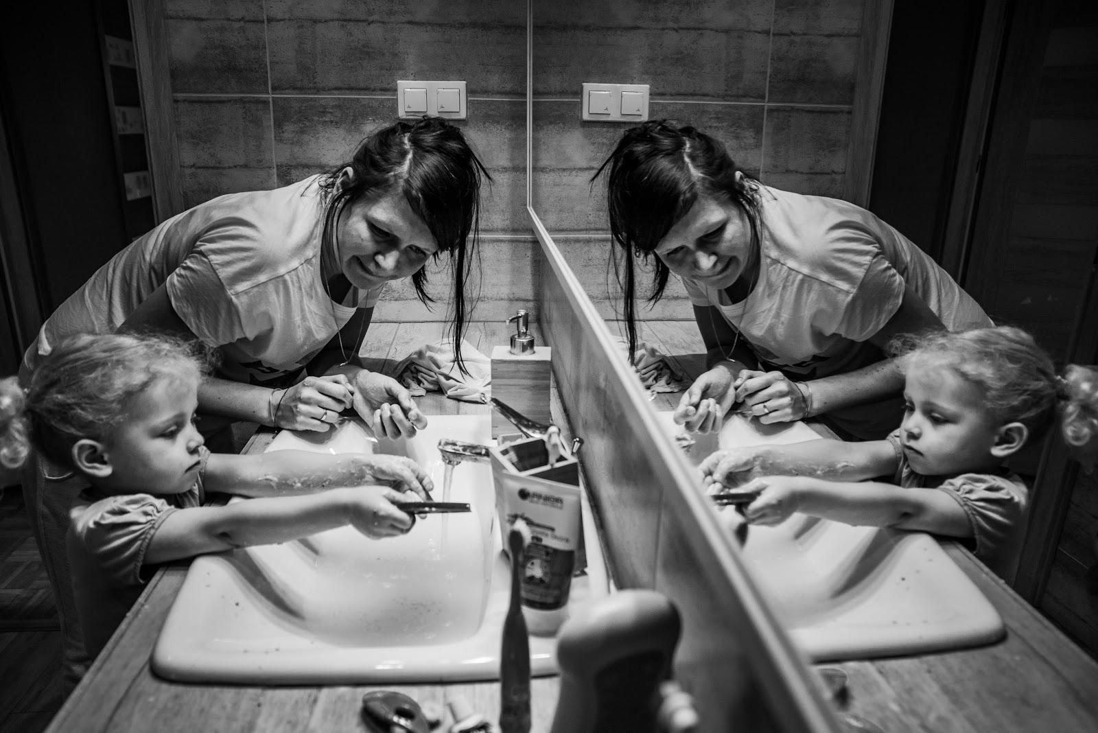 Czarno-biała fotografia. Na zdjęciu widoczna mama z dzieckiem stojące w łazience przy umywalce. Mama nachylona do dziecka przygląda się, dziecko pod odkręconym kranem trzyma jakiś przedmiot. W kadrze przedstawiona scena odbija się w lustrze.