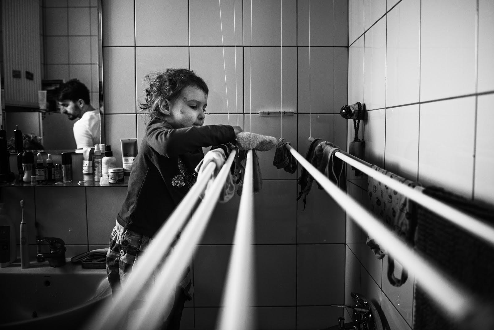 Czarno-biała fotografia. Na niej widoczne kilkuletnie dziecko w łazience rozwieszające pranie na szuszarce wiszącej nad wanną. Z lewej strony kadru widoczne odbicie ojca w lustrze na umywalką.