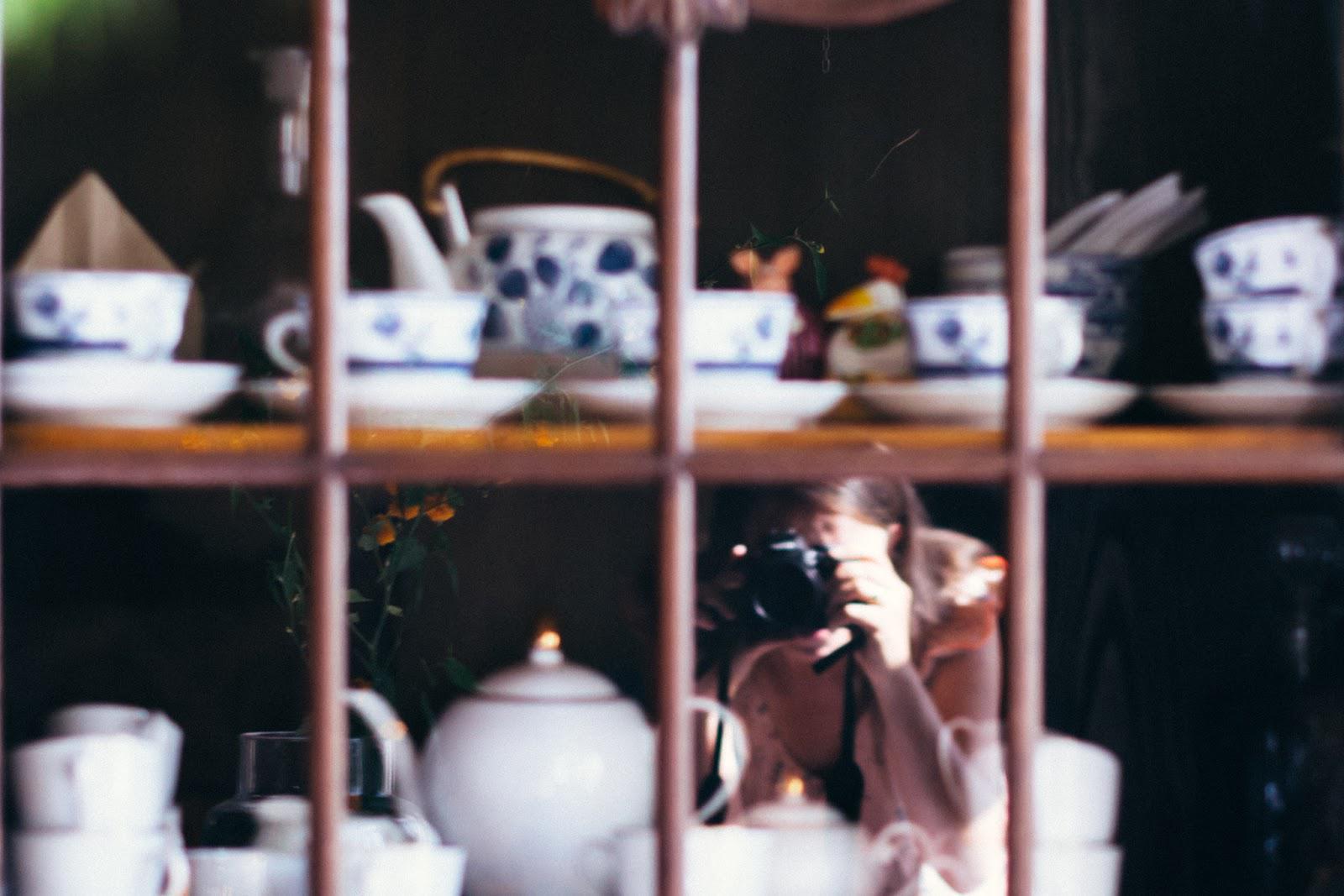 Kolorowa fotografia fragmentu szafki z ustawionym wewnątrz zestawem porcelany. Widoczne są białe filiżanki z niebieskimi zdobieniami z dzbankiem oraz poniżej drugi zestaw - białego koloru. W szybie szafki odbija się fotografka robiąca to zdjęcie.