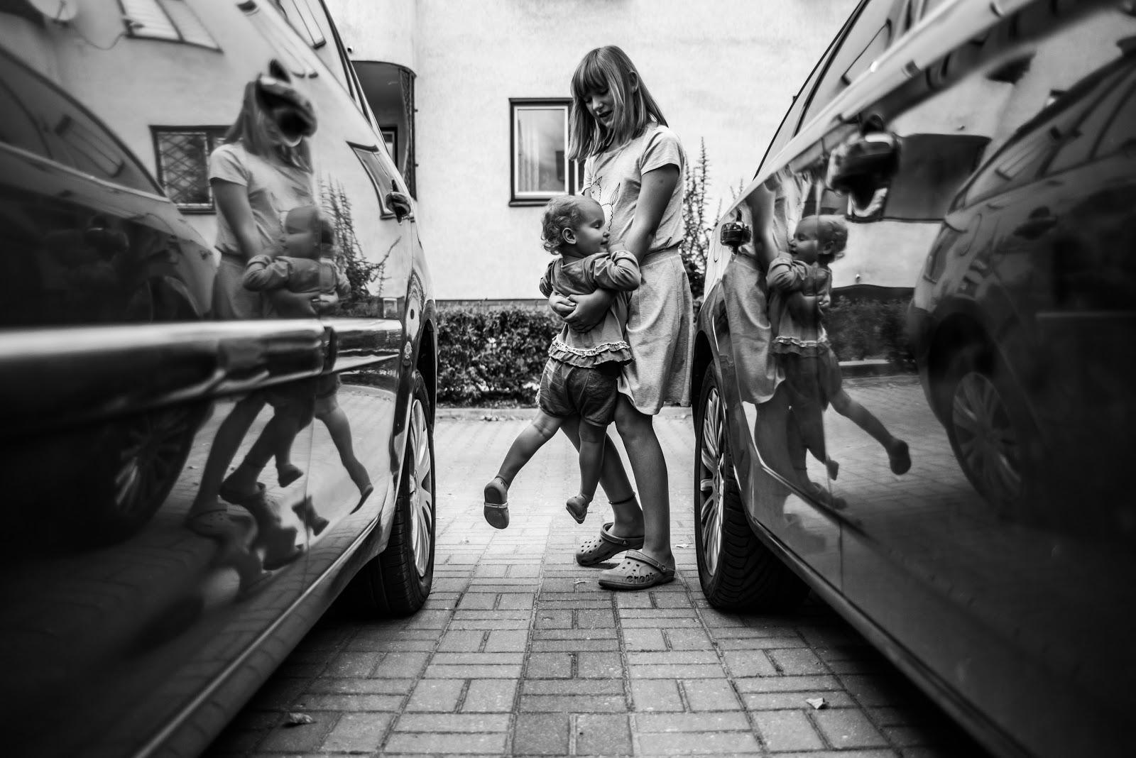 Czarno-biała fotografia mamy bawiącej się z dzieckiem. Mama unosi dziecko delikatnie nad ziemią. Obie osoby znajdują się pomiędzy samochodami, co powoduje odbicia postaci w karoseriach samochodów.